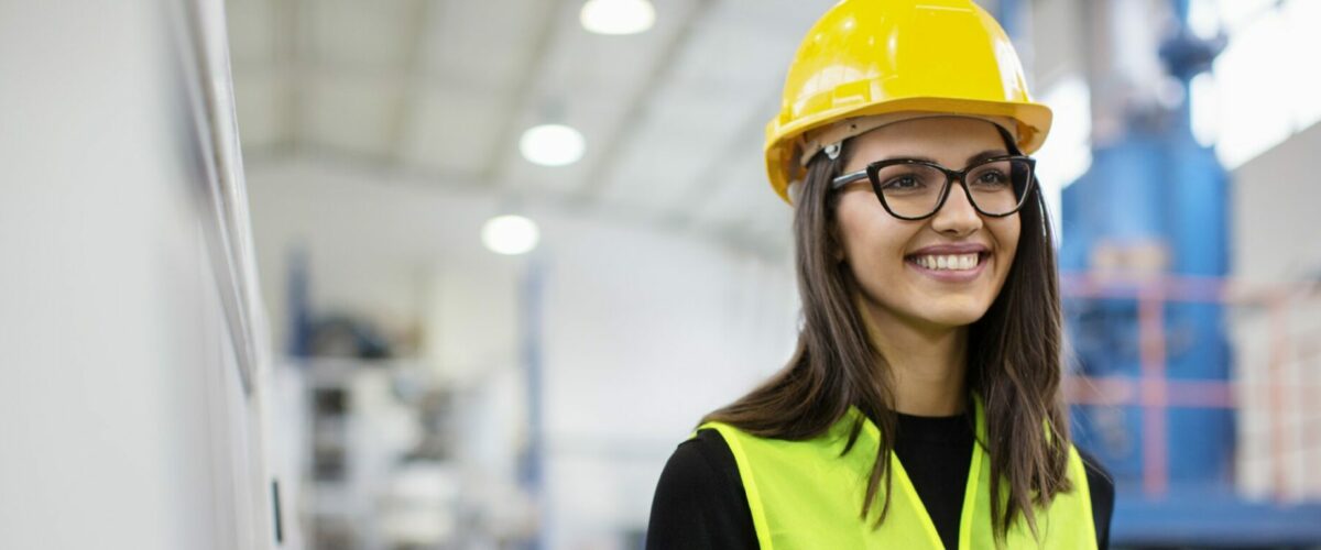 Schönes Mädchen in der Fabrik in einer Weste und einem Helm, das lächelt und einen Händedruck macht.